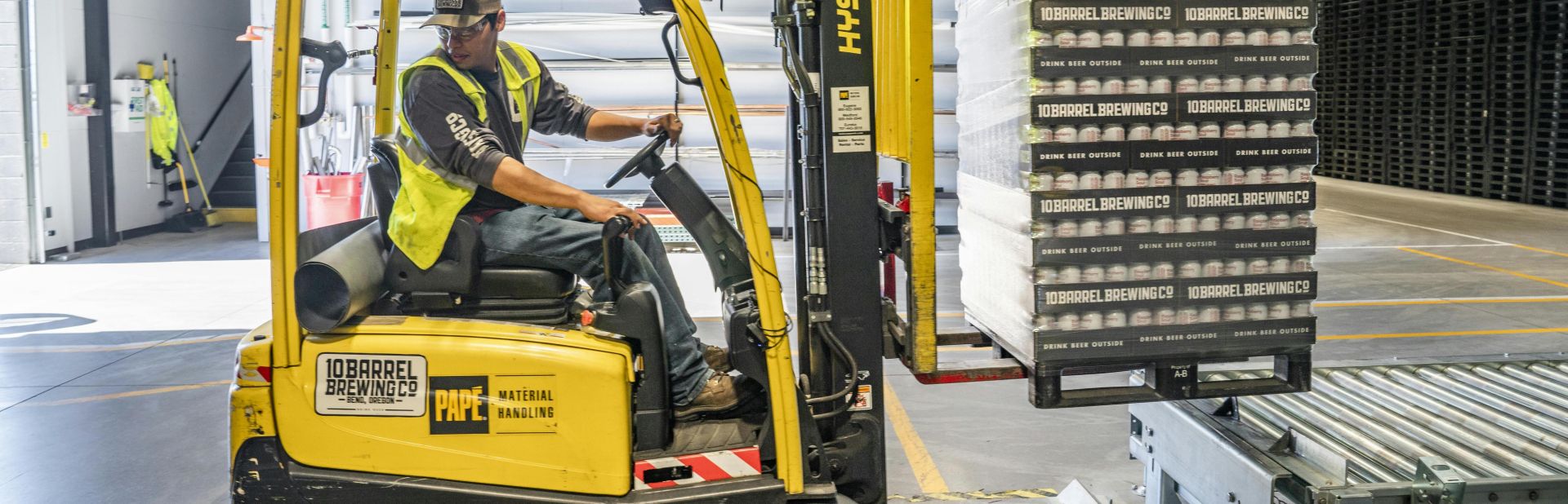A warehouse worker maneuvers a forklift to transport crates for brewing company storage.