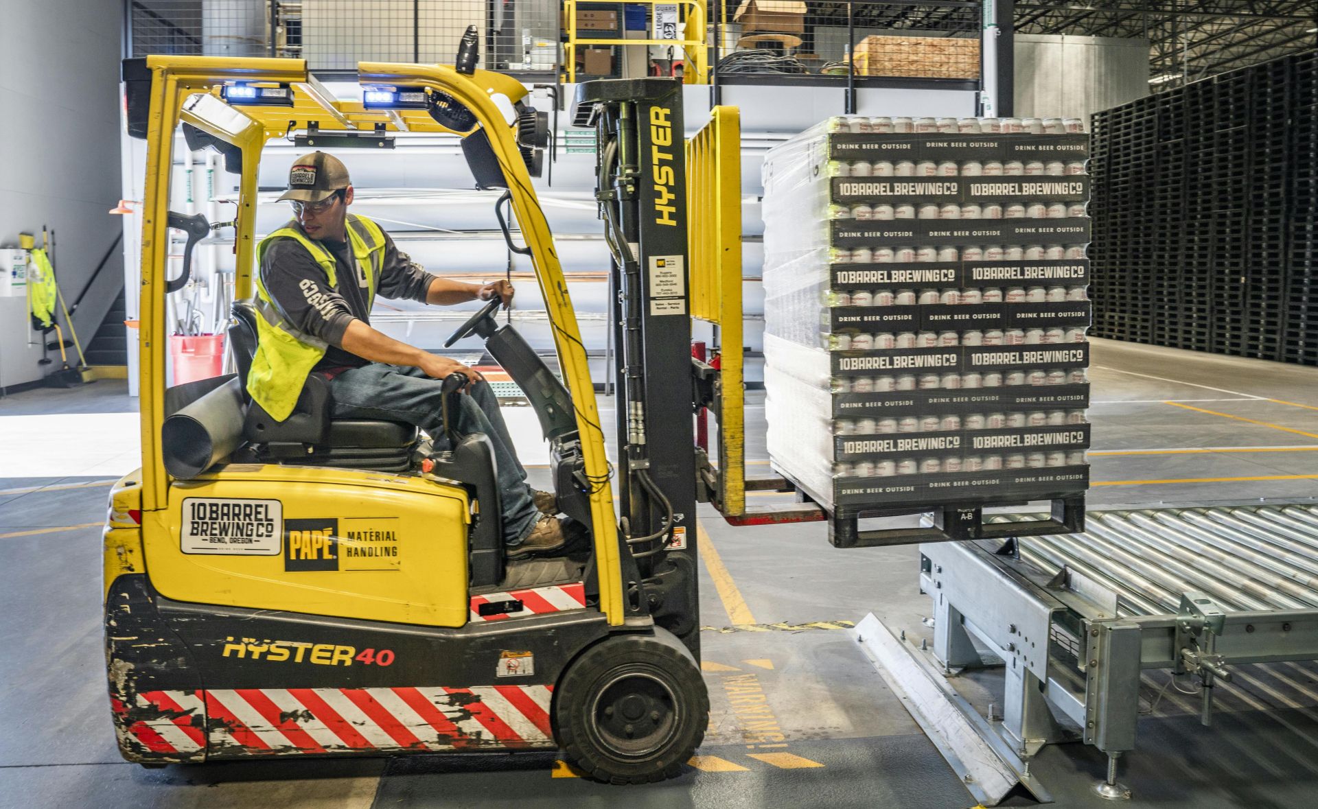 A warehouse worker maneuvers a forklift to transport crates for brewing company storage.