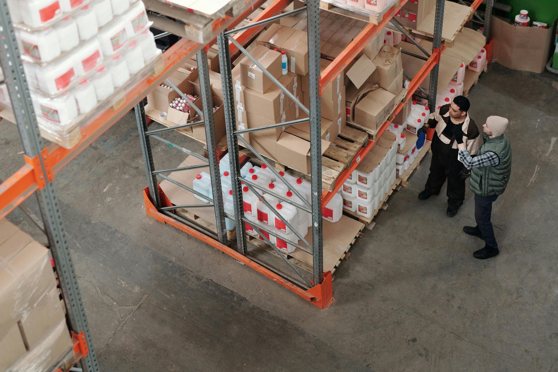 Two warehouse workers discussing logistics beside shelving with boxes and containers.