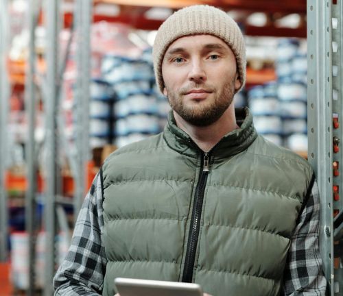 A man wearing a beanie and vest holding a tablet in a warehouse setting.